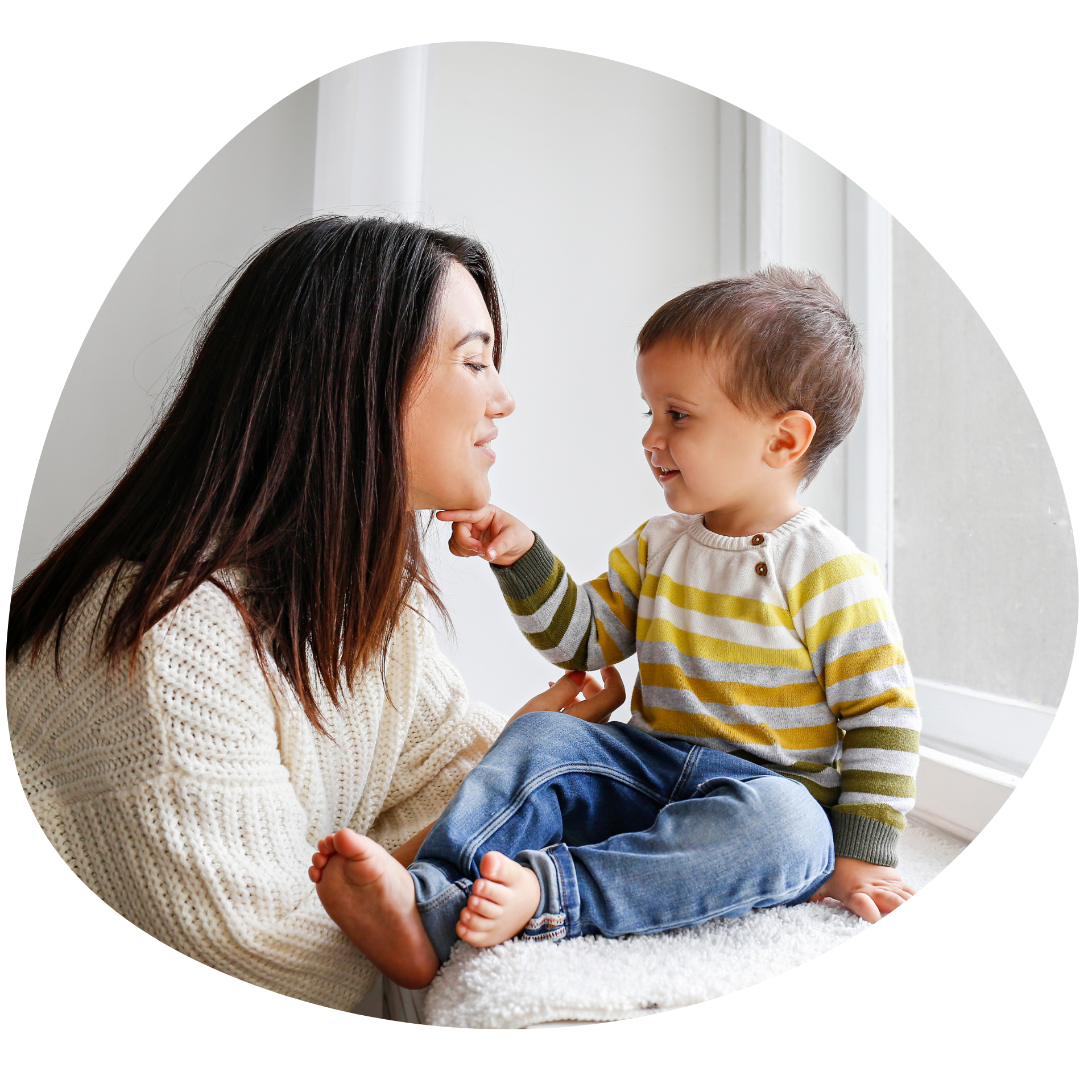Woman and toddler smile at each other, window seat. Child touches her chin, wearing jeans and striped shirt.