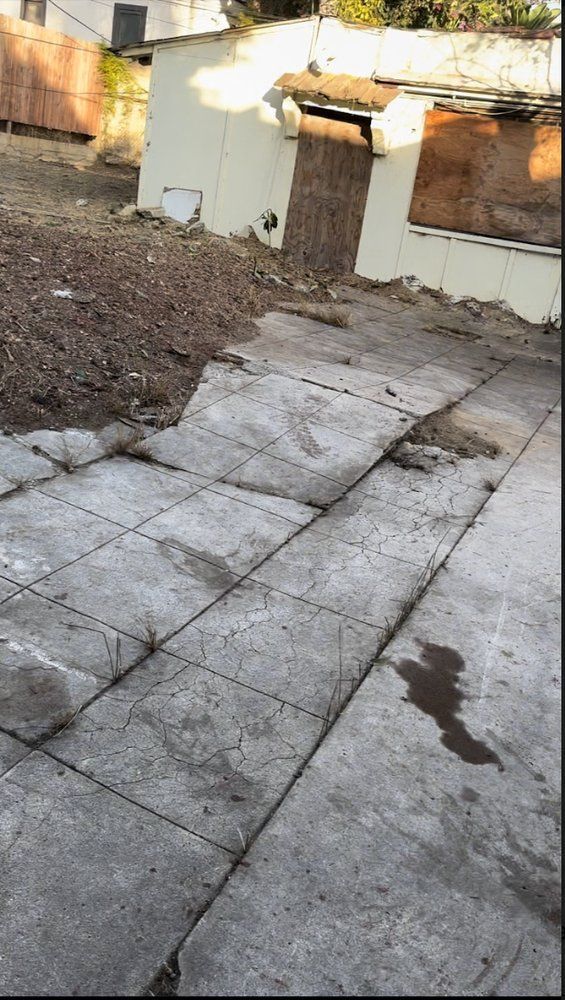 Concrete patio and dirt pile in a yard, partially visible building with boarded up windows.