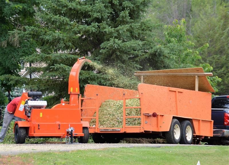 Orange wood chipper and trailer being loaded with branches; man works on the machine in front of trees.