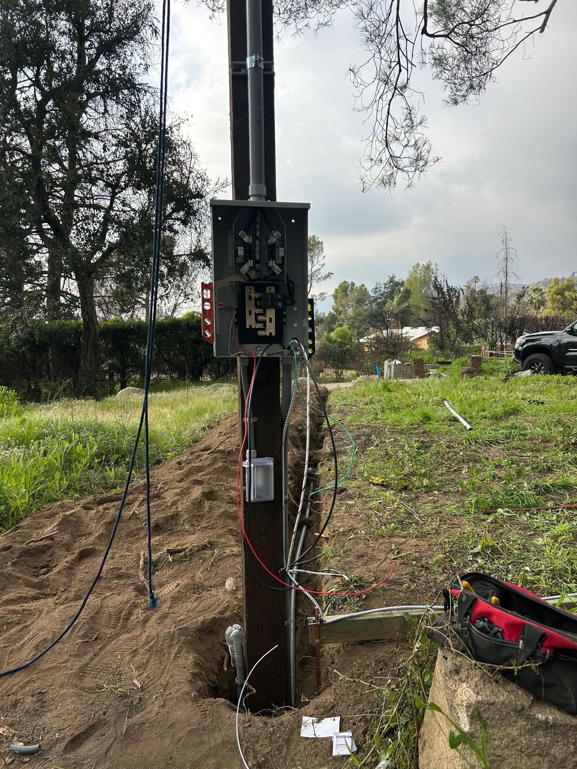 Utility electrical box on a wooden pole in a field, with wires and tools nearby under a cloudy sky