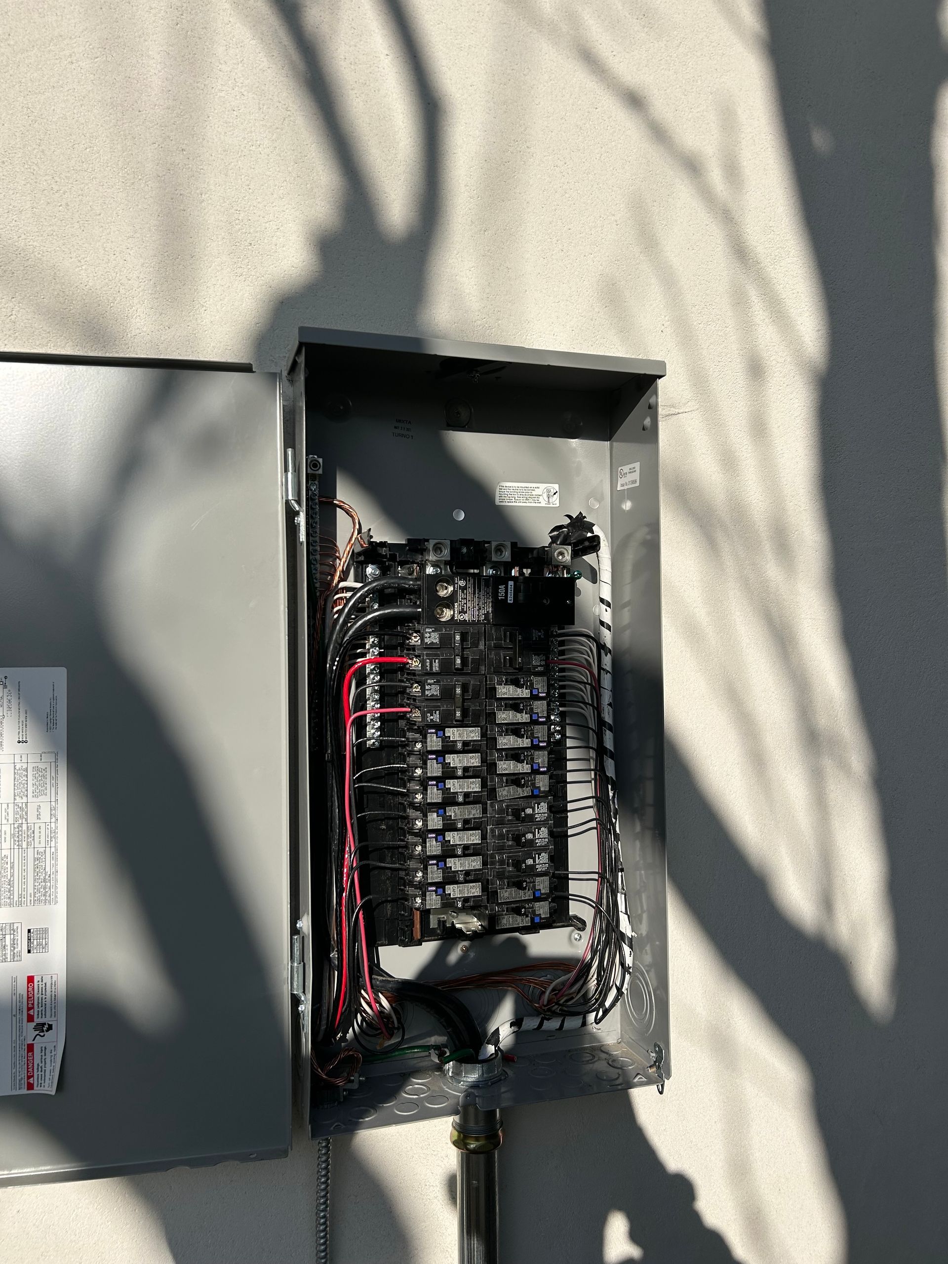 Open electrical breaker panel with exposed wiring on a beige exterior wall.