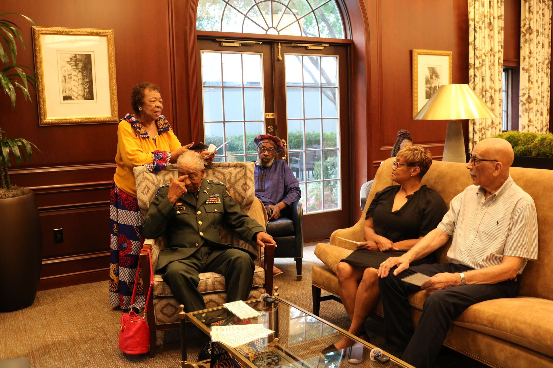 A group of people are seated and standing in a well-lit living room, engaged in conversation.