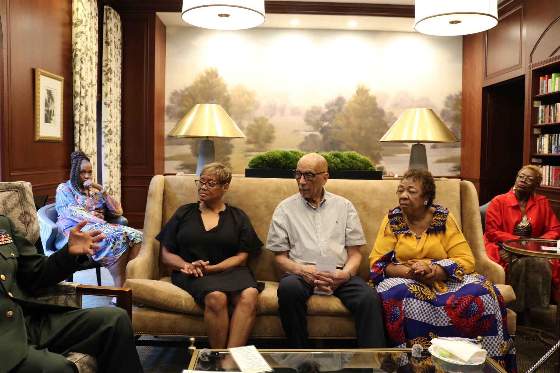 People seated on a couch, listening to a person speaking. Indoors, neutral tones.