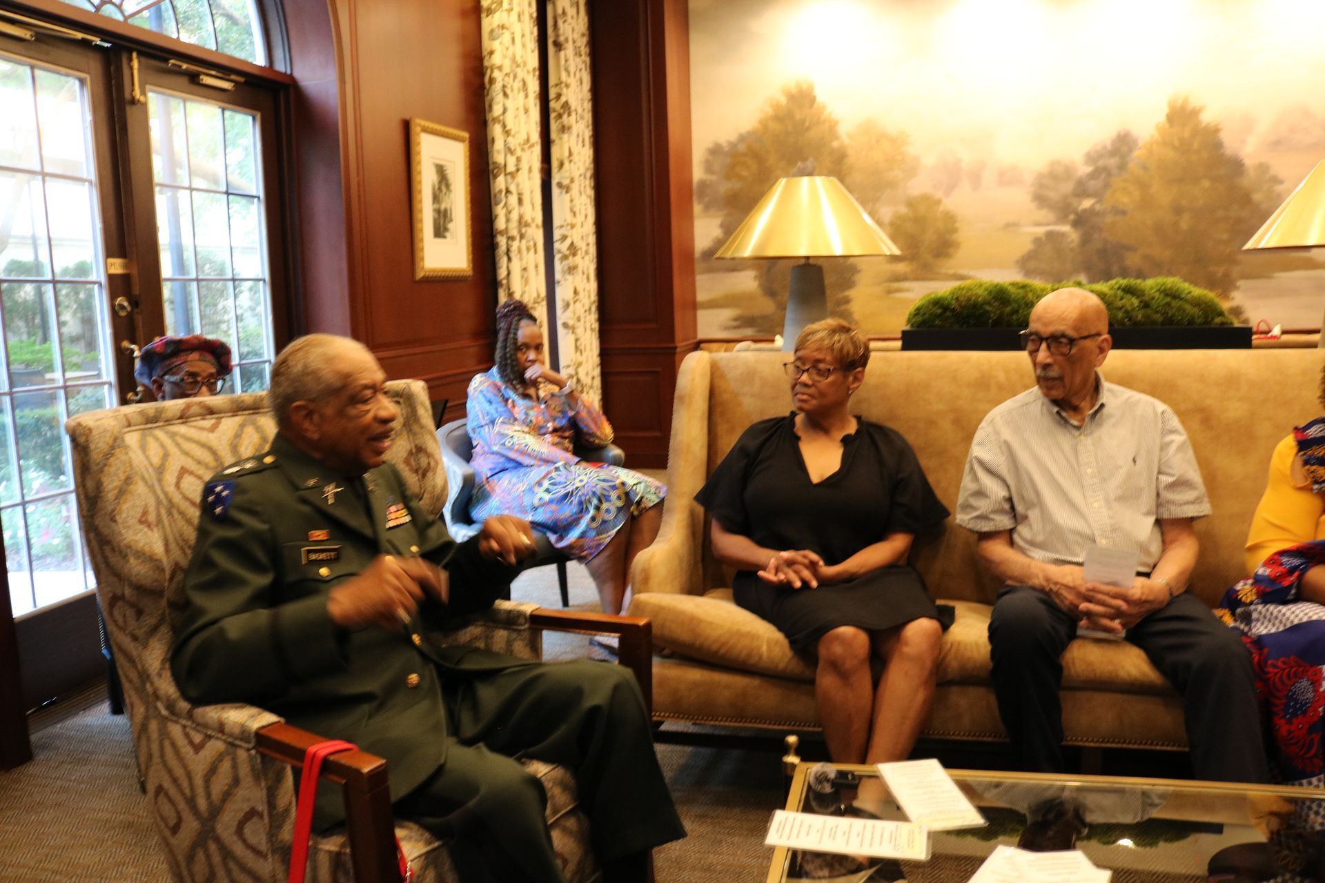 Group of people in a room, some seated, in conversation. One in military uniform. Interior setting.