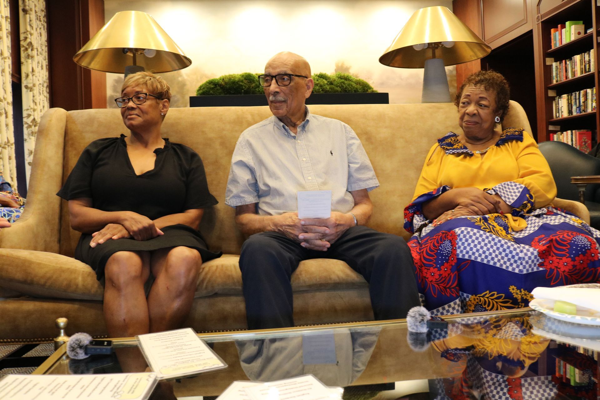 Three people sitting on a sofa indoors. One holds papers, others look forward. Yellow, blue, and black fabrics.