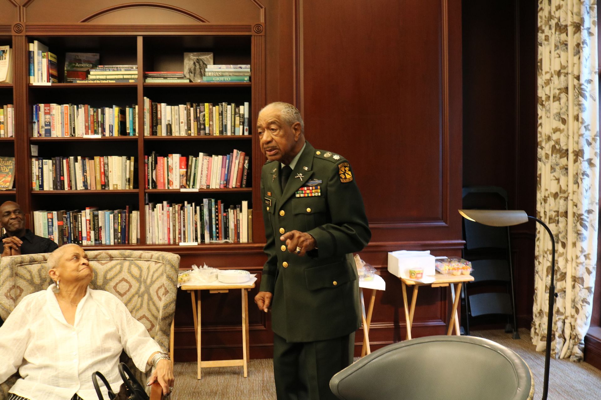 Man in military uniform speaks to seated person, bookshelves in background.