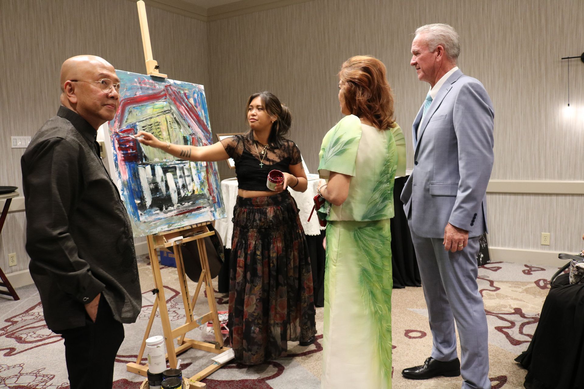People viewing a woman painting at an event; painting on an easel, other people nearby.