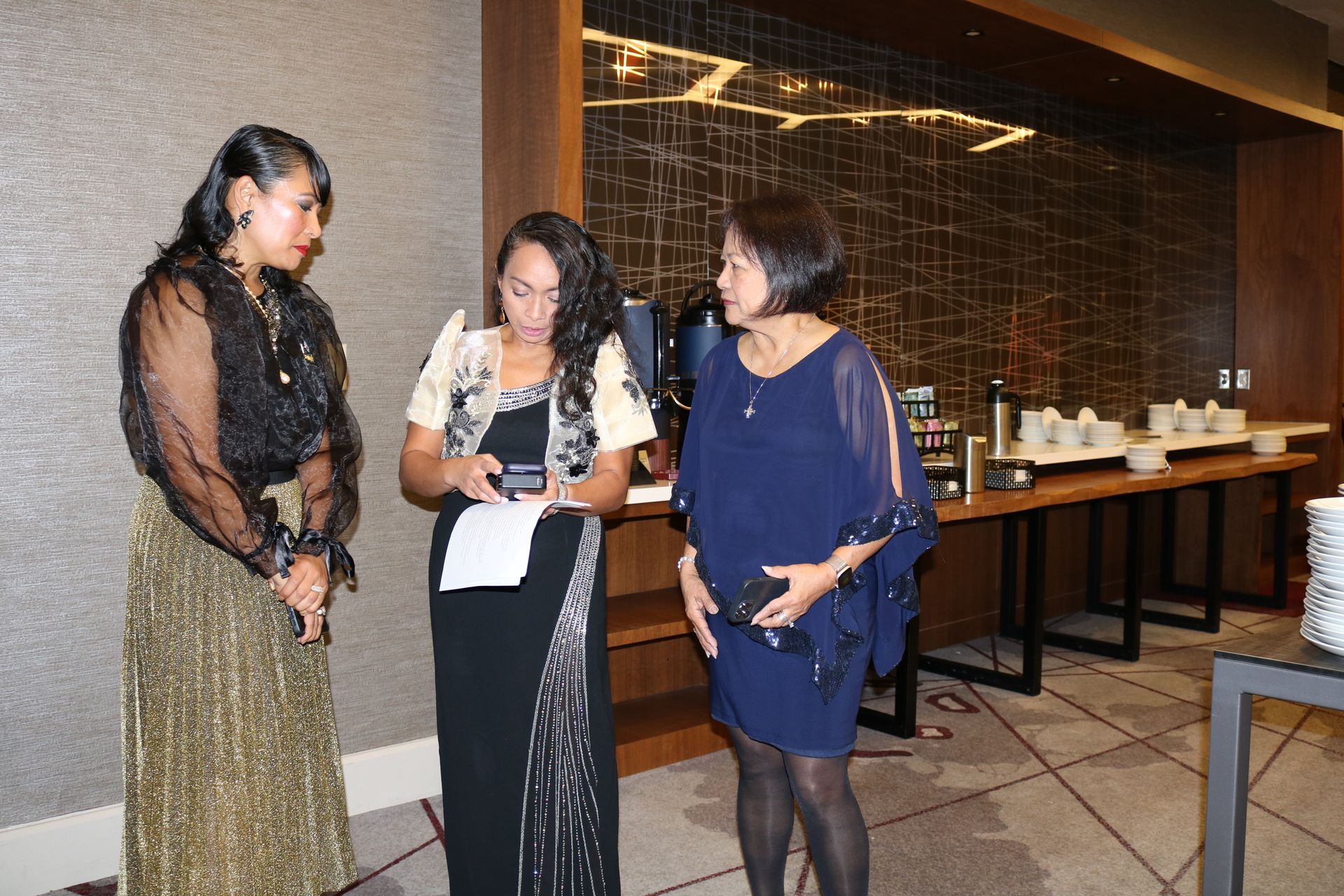 Three women in formal attire conversing near a buffet table. Two women look at a paper.