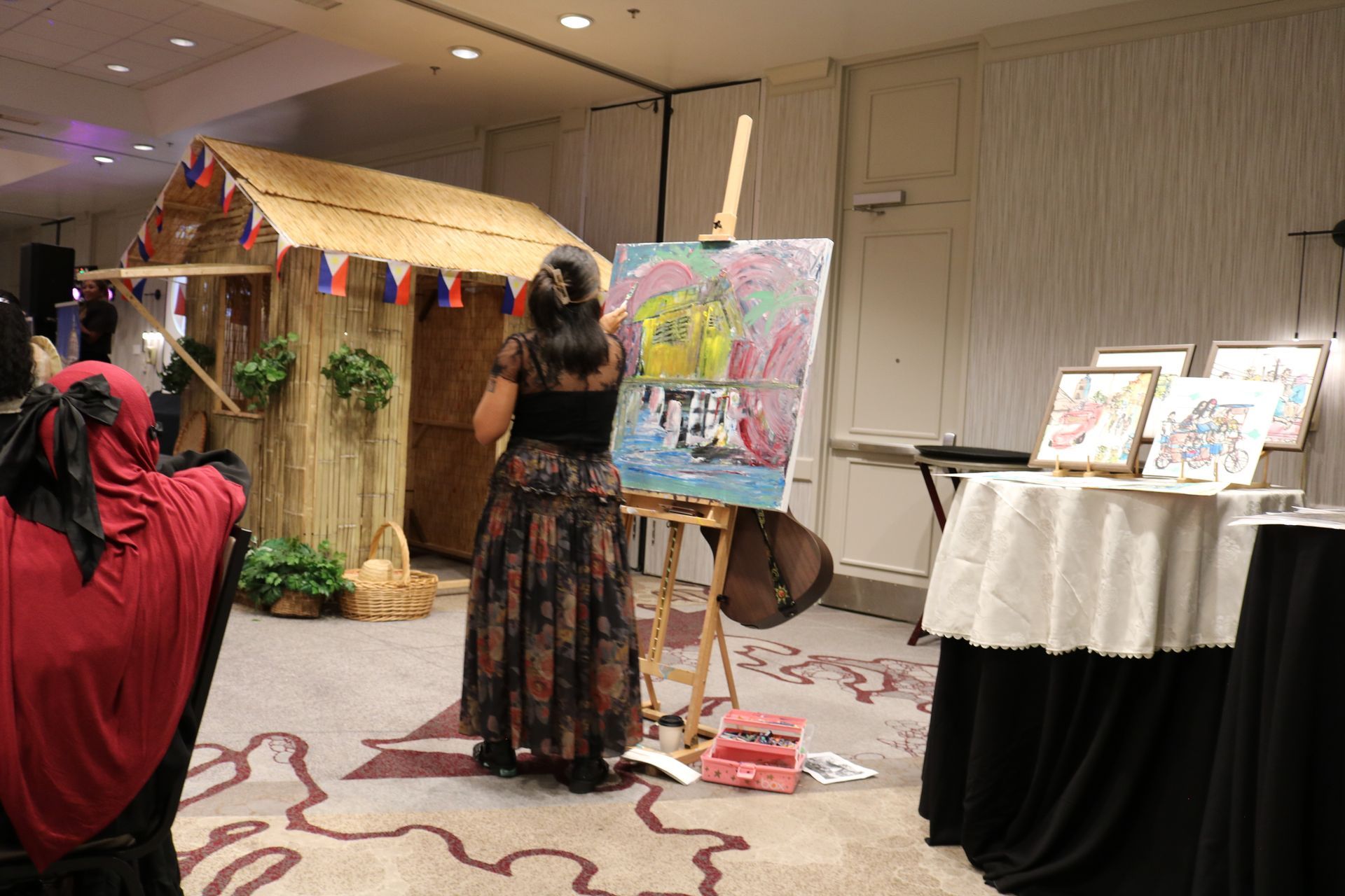 Woman painting at an easel near a bamboo hut, other paintings on display at a formal event.