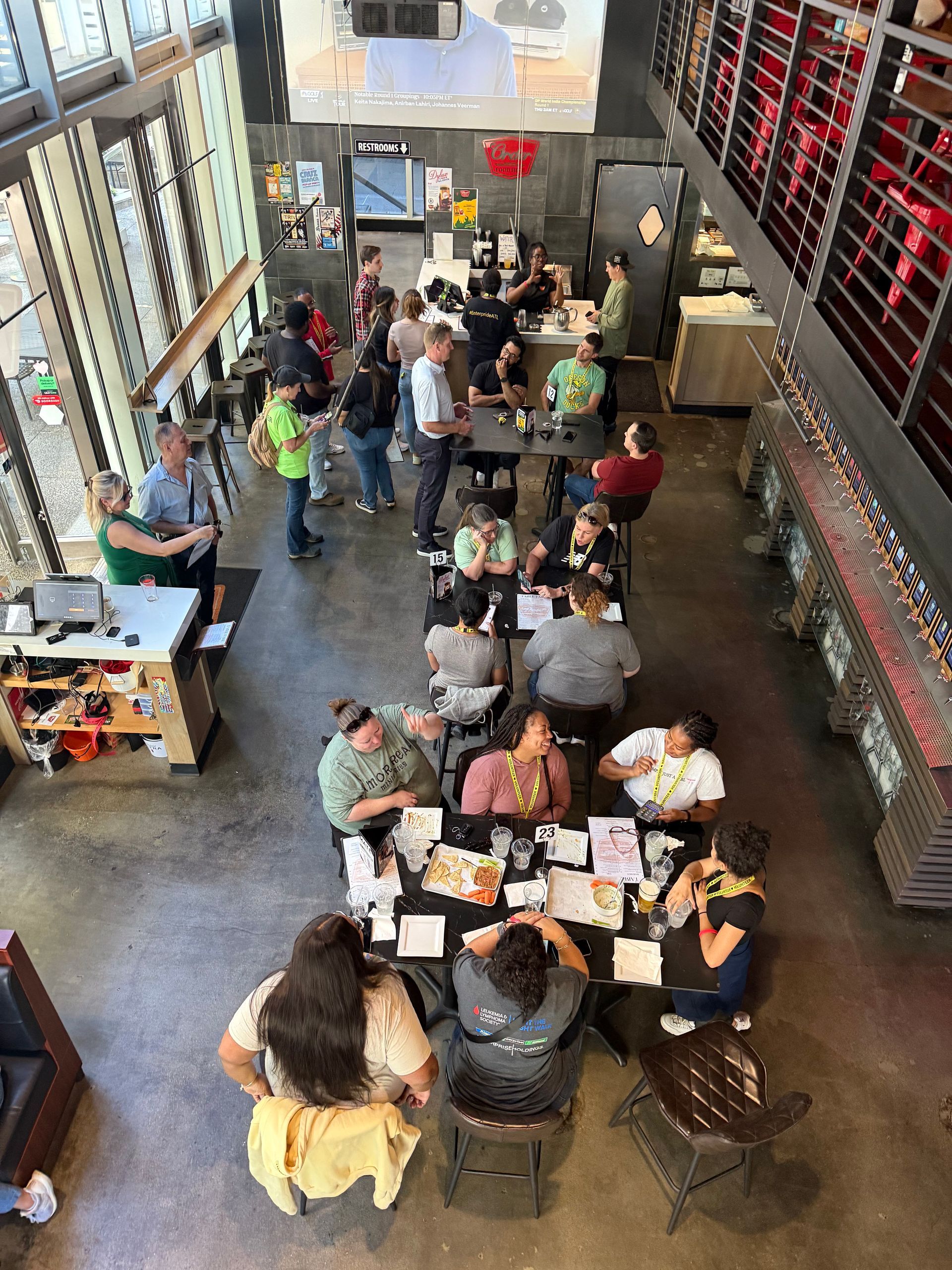 Group of people at tables in a workshop, some seated and working. Overhead view, bright, industrial setting.