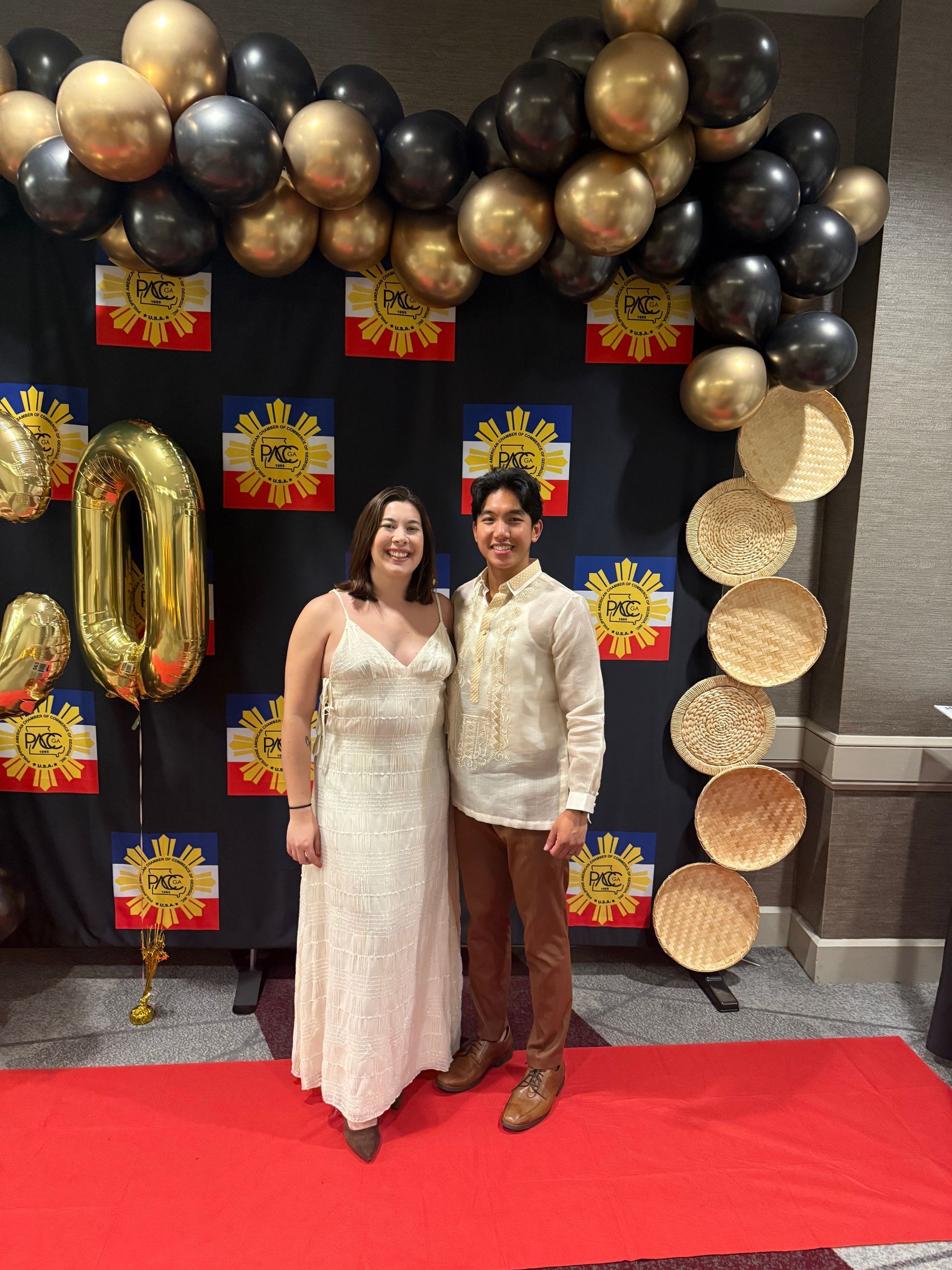 Two people pose in front of a backdrop with balloons and Philippine flags. The woman wears a white dress; the man, a brown shirt and pants.