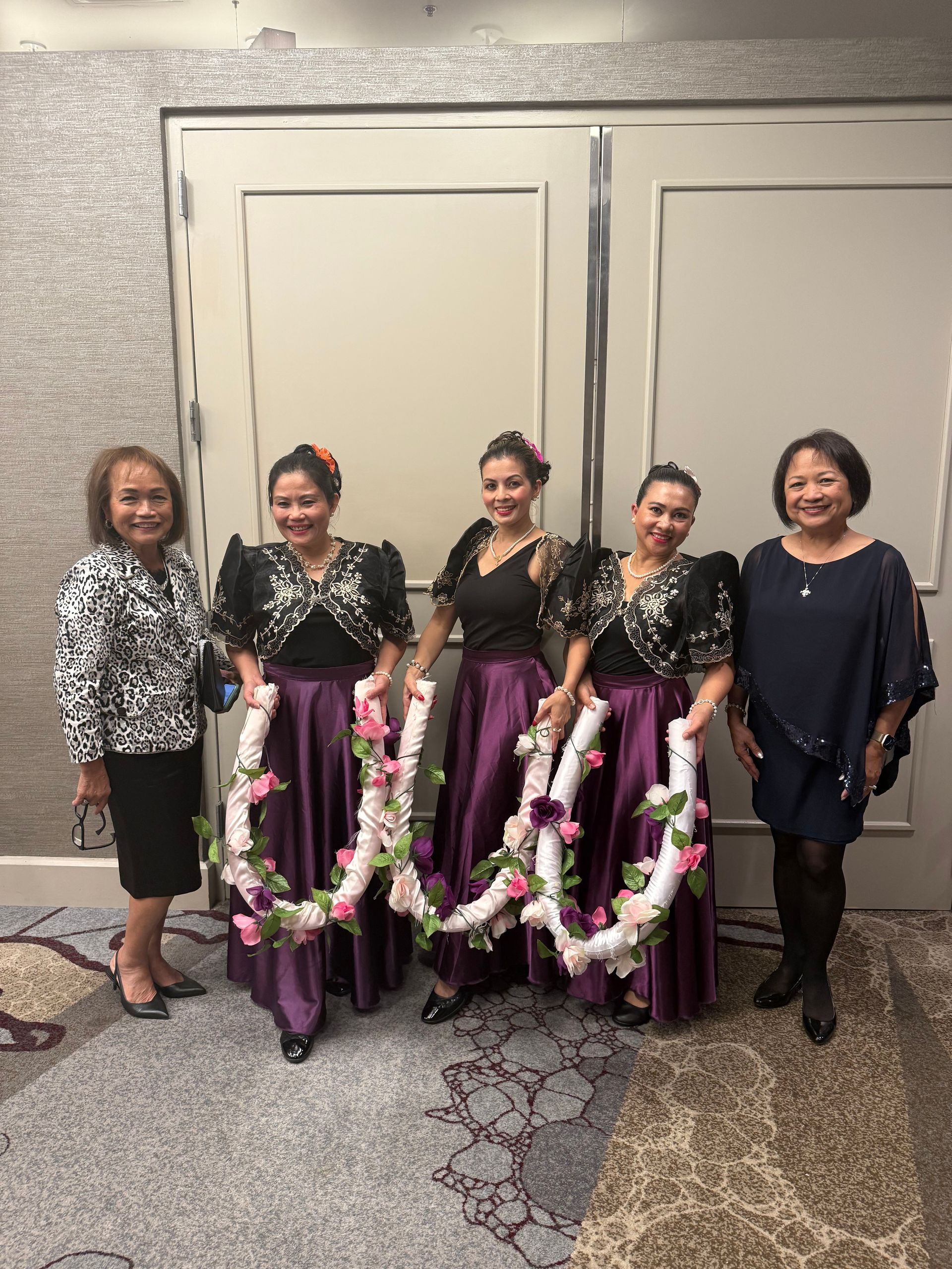 Five women stand in front of double doors; three in purple skirts and black tops hold floral garlands.