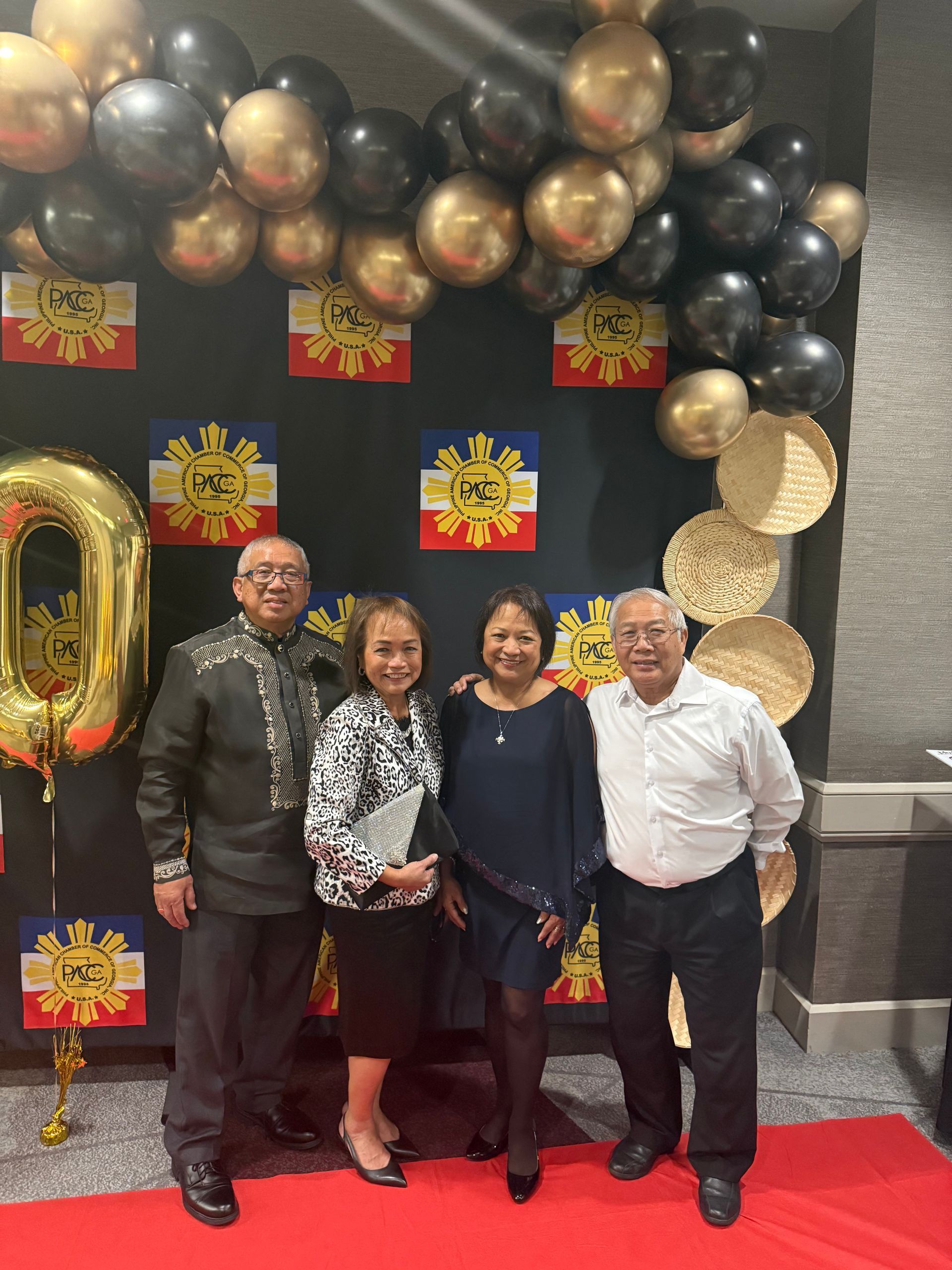 Four people pose in front of a backdrop with gold and black balloons, Philippine flags.