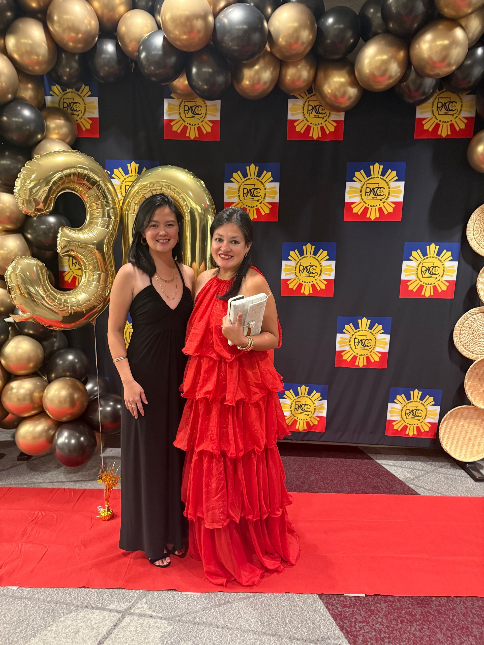 Two women pose on a red carpet in front of a backdrop with balloons and Philippine flags. The woman on the left wears a black dress. The woman on the right wears a red dress.