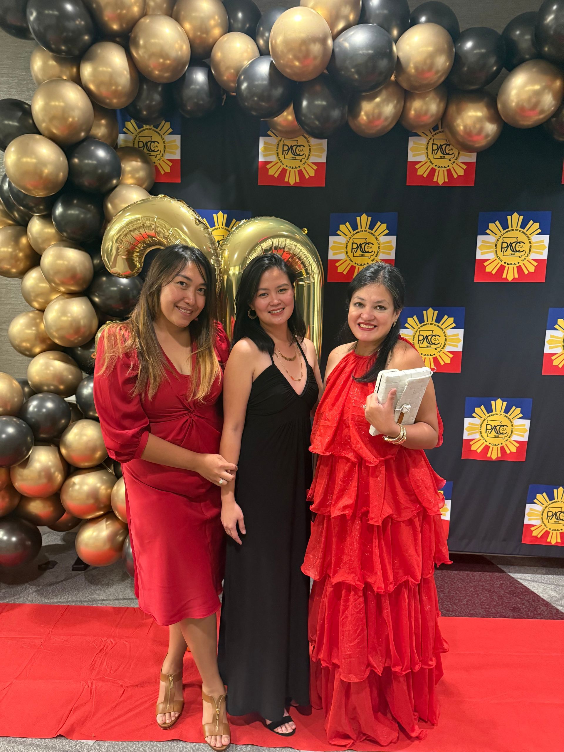 Three women posing on a red carpet in front of balloons and Philippine flags. Two wear red dresses, one black.