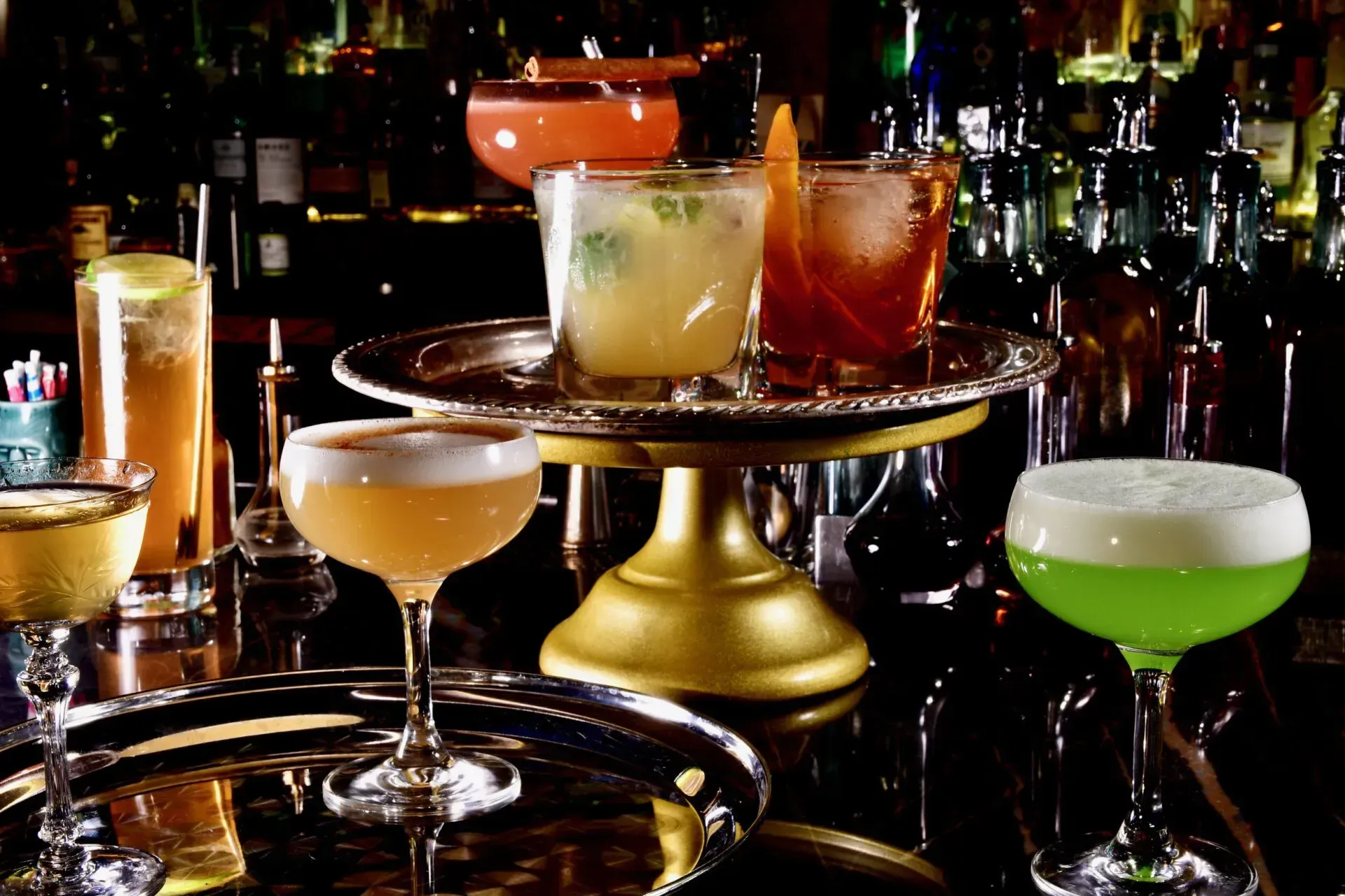 Cocktails on a bar, various colors, some with foam. Silver trays, bottles in background.