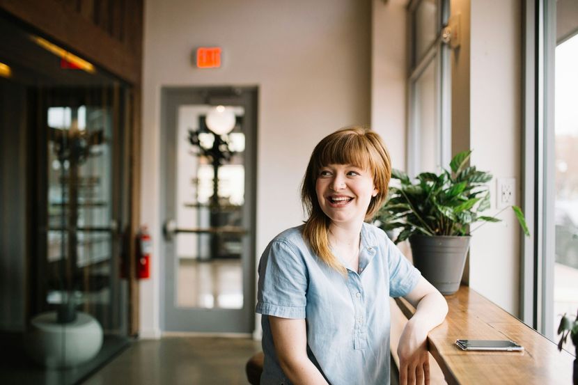 A woman is sitting at a table in front of a window and smiling.