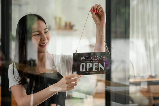 A woman is holding a welcome open sign in front of a glass door.