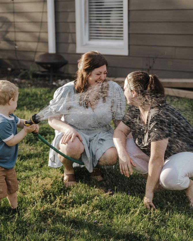 Two women and a baby are playing with a hose in the backyard.