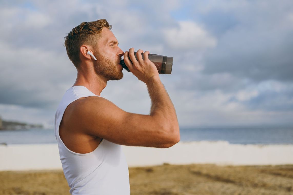 Man drinking from a bottle on a grassy shoreline under cloudy sky