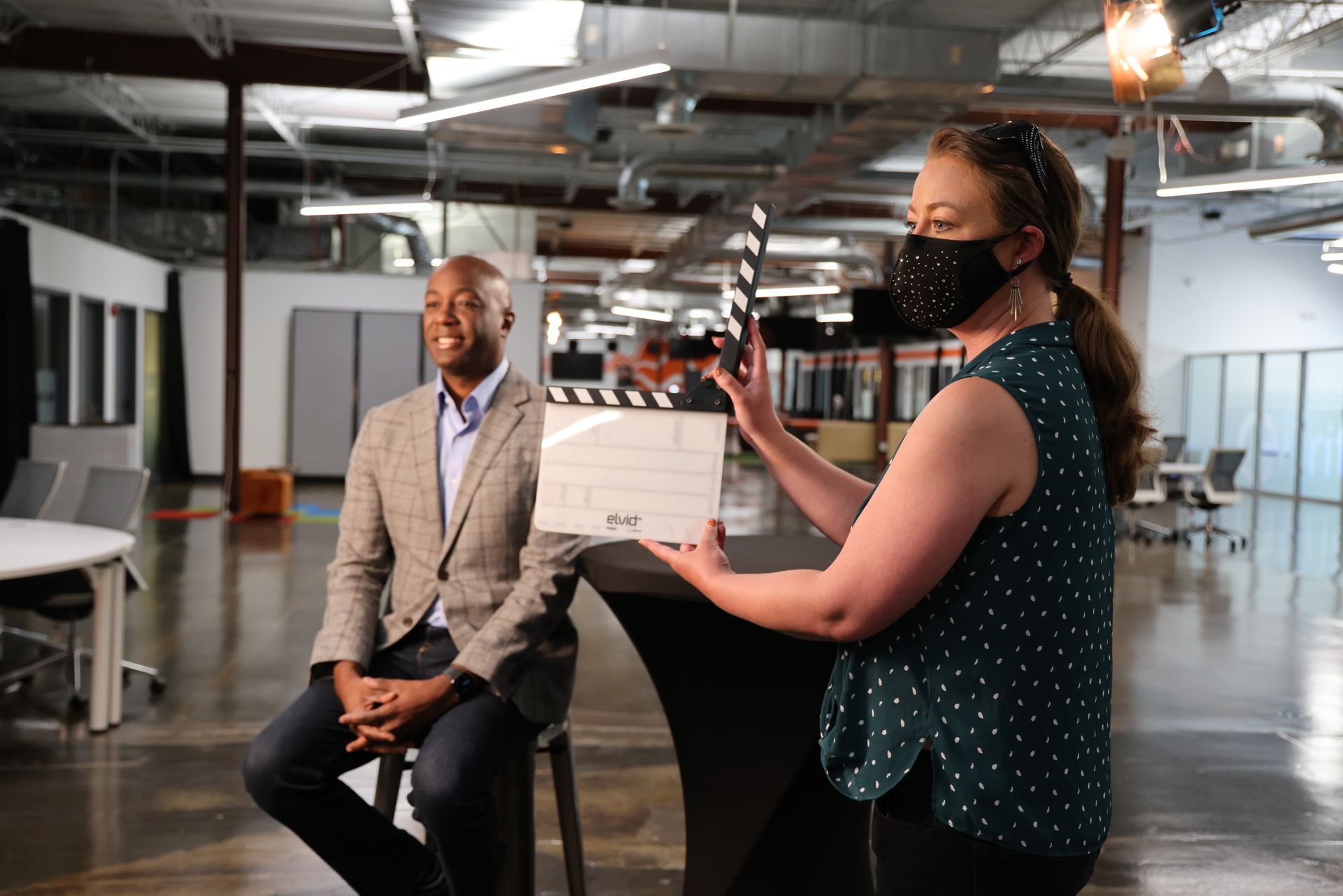 A woman wearing a mask is holding a clapper board in front of a man sitting on a stool.