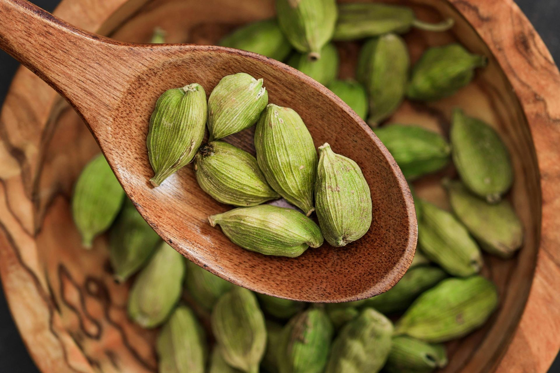 A wooden spoon filled with green cardamom seeds next to a bowl of cardamom seeds.