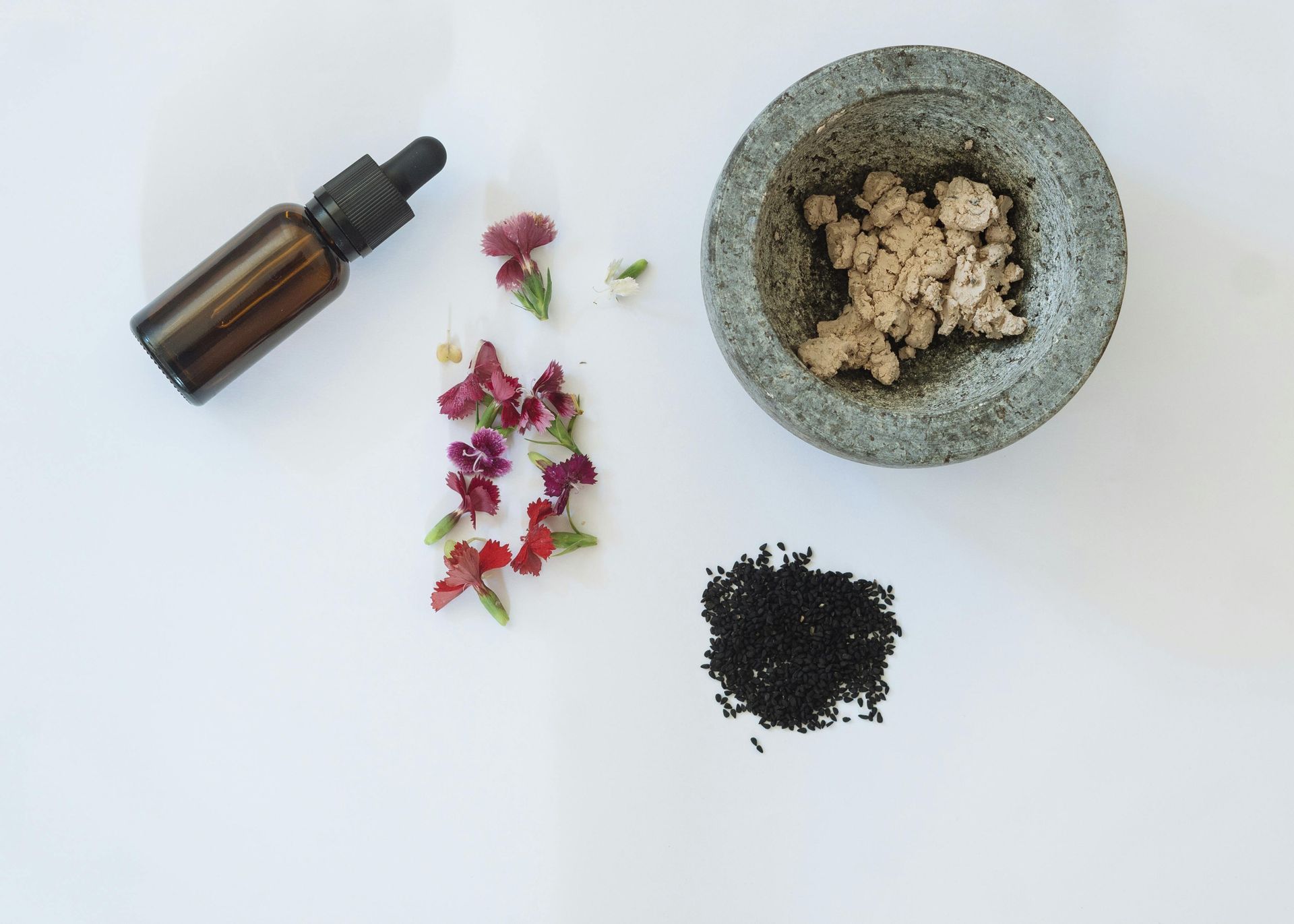 A mortar and pestle filled with seeds and flowers next to a bottle of essential oil.