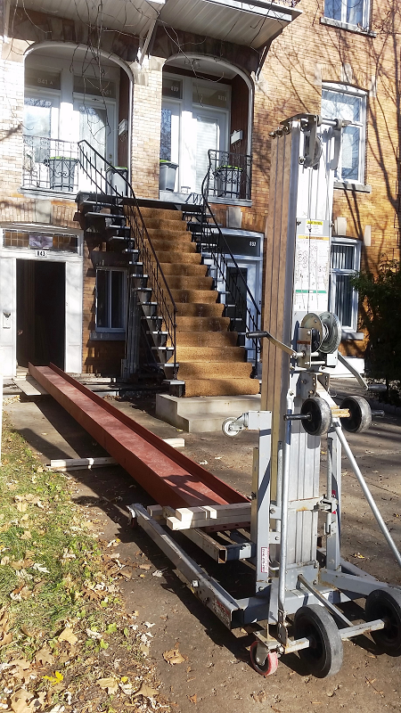 Une grande poutre métallique est soulevée par une machine devant un bâtiment.