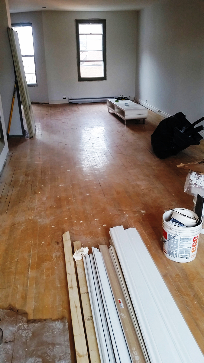 Un salon avec du parquet et un seau de peinture au sol.