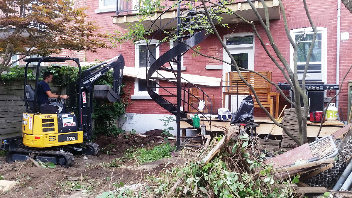 Un homme conduit une petite pelle devant un bâtiment en brique.