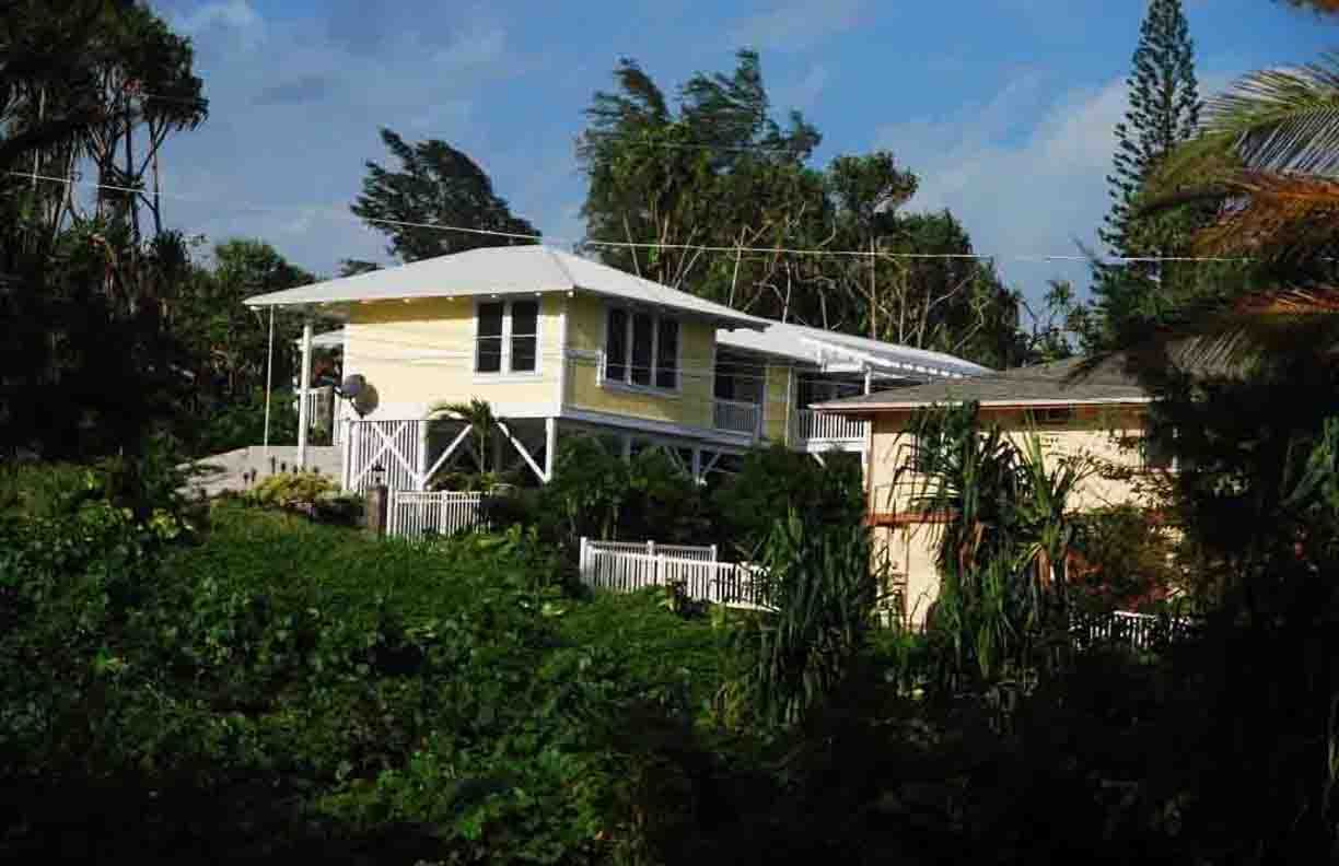 Yellow house on a hill, surrounded by lush green trees under a partly cloudy sky.