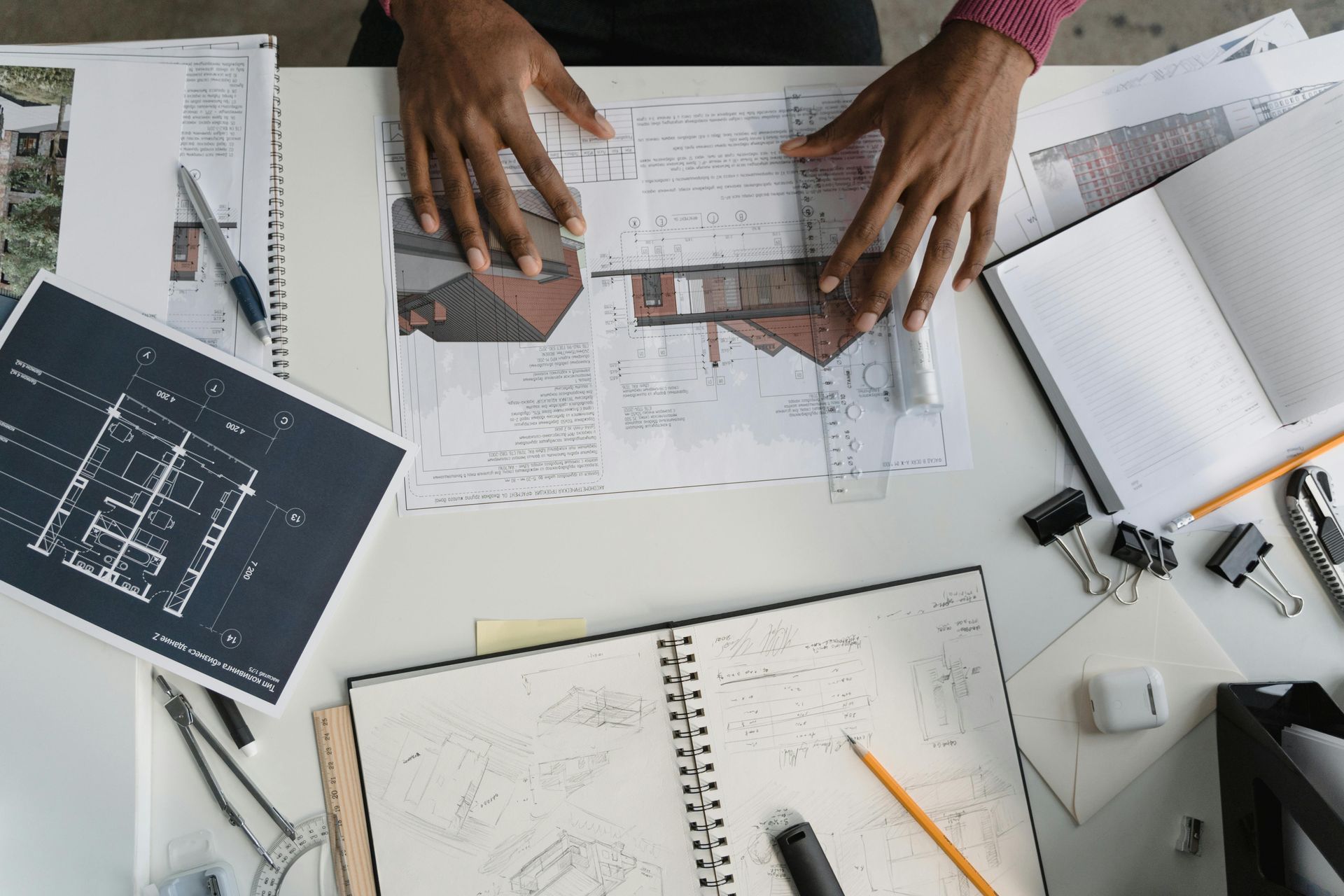 An overhead view of hands working on architectural blueprints and sketches at a desk with drafting tools and notebooks.