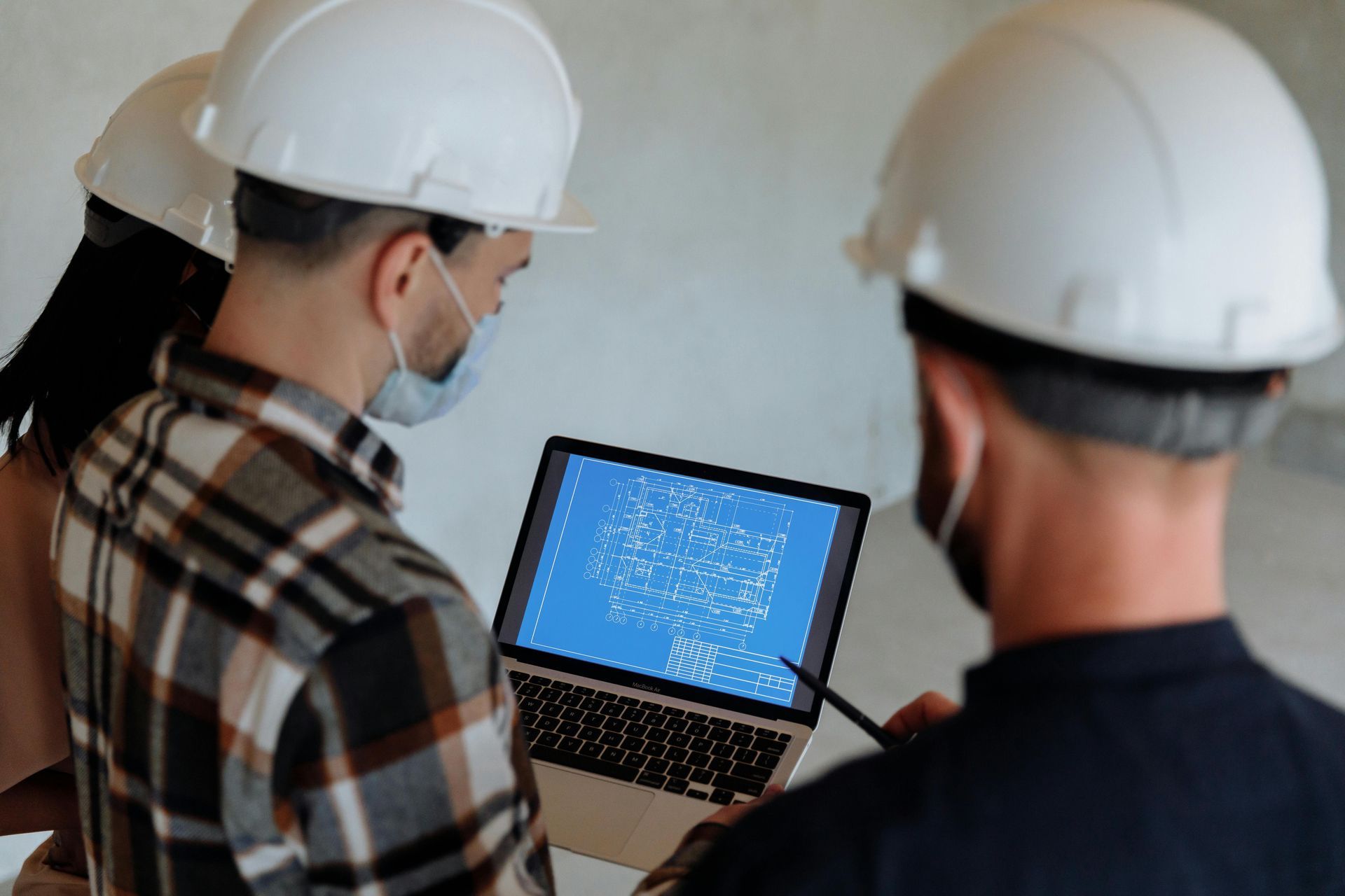Three people in hard hats and masks looking at a laptop with blueprints.