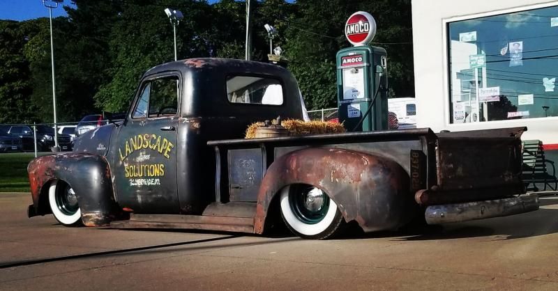 Rusty black vintage pickup truck with whitewall tires parked at a gas station.