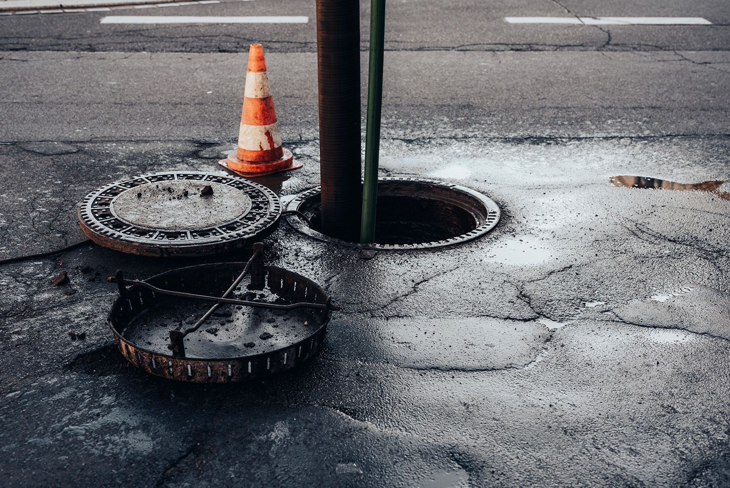 A manhole cover is open on the side of the road next to a traffic cone.