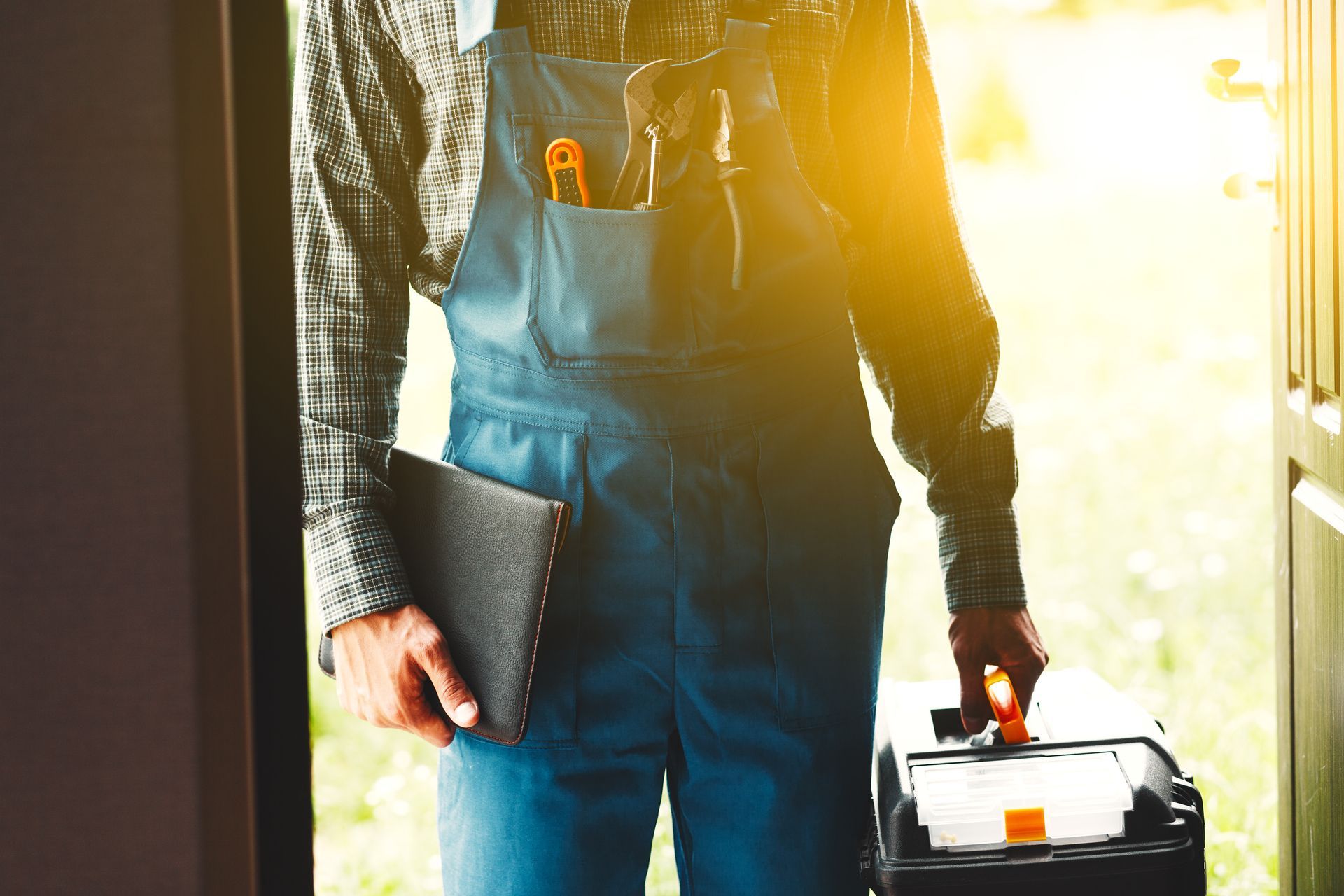 Worker in blue overalls holds toolbox and tablet in sunlit doorway, tools in pocket.