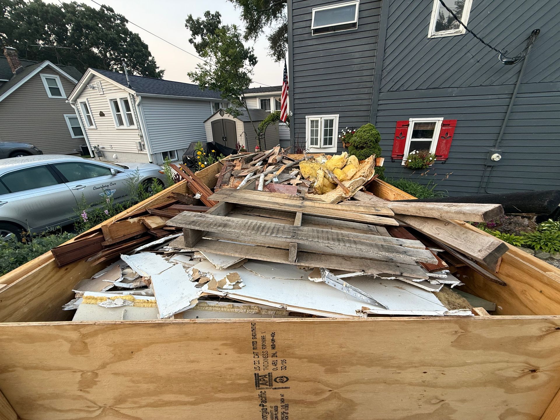 A wooden dumpster filled with construction debris in front of houses.