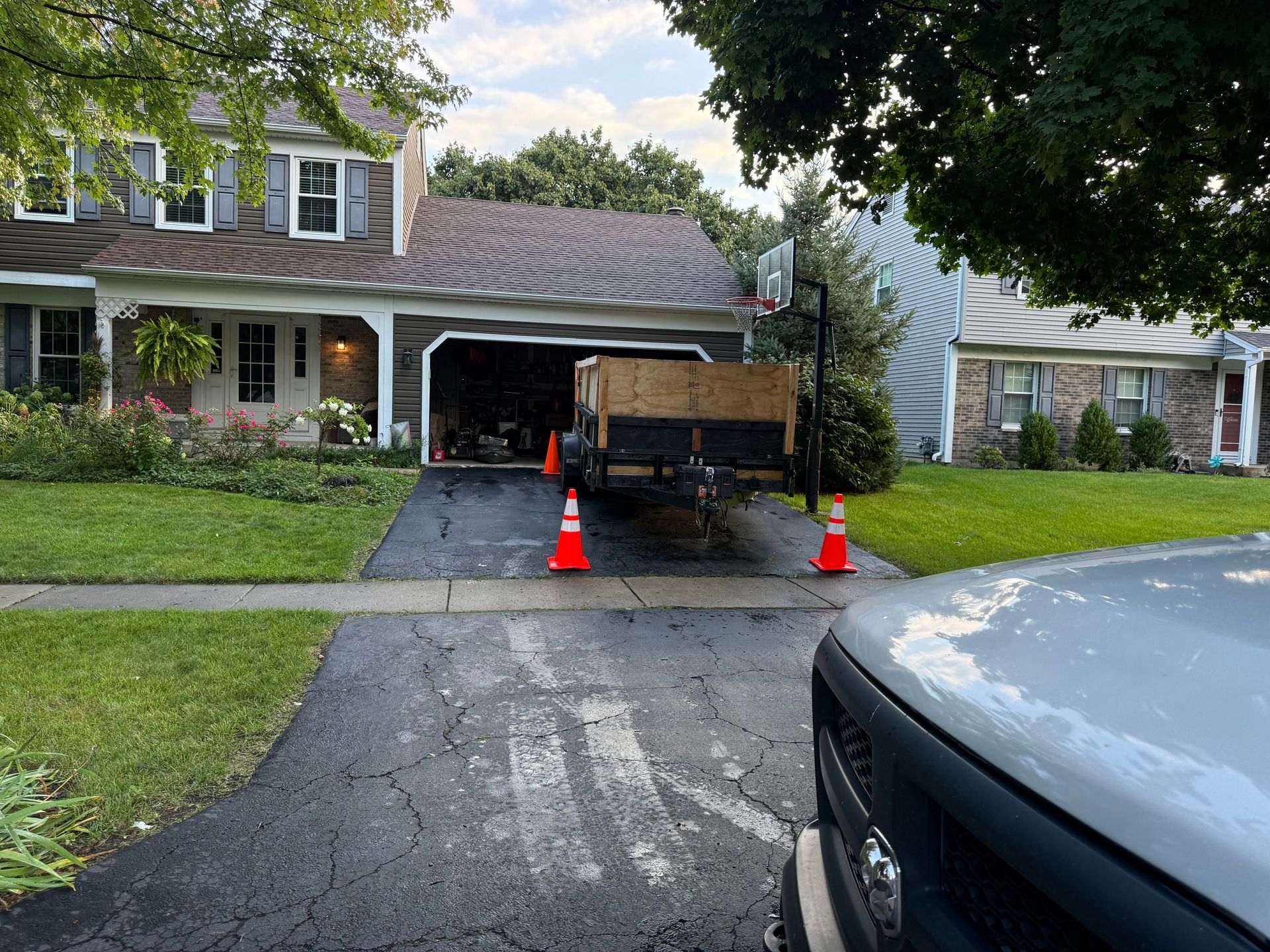 A black paved driveway with a trailer, orange cones, and a two-story house.