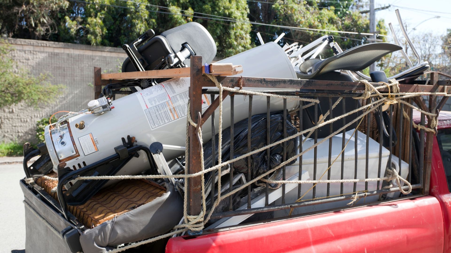 Red pickup truck bed overloaded with scrap metal, secured with rope, on a street.