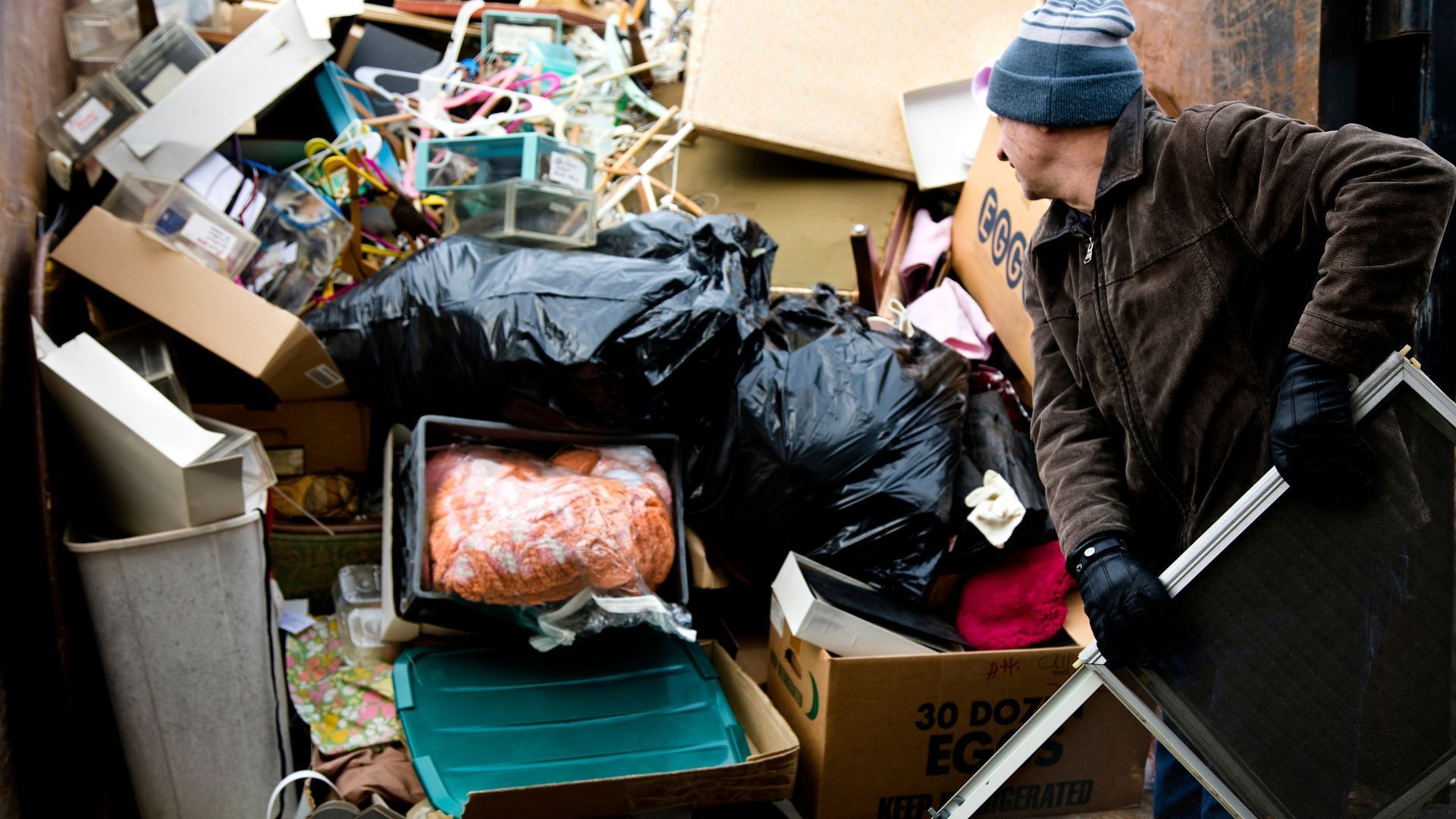 Man rummages through dumpster full of trash; boxes, bags, and other items.
