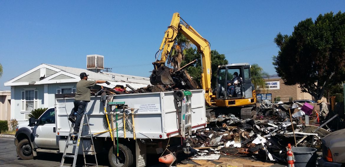A yellow excavator loads debris into a dump truck; a man stands on a ladder assisting.