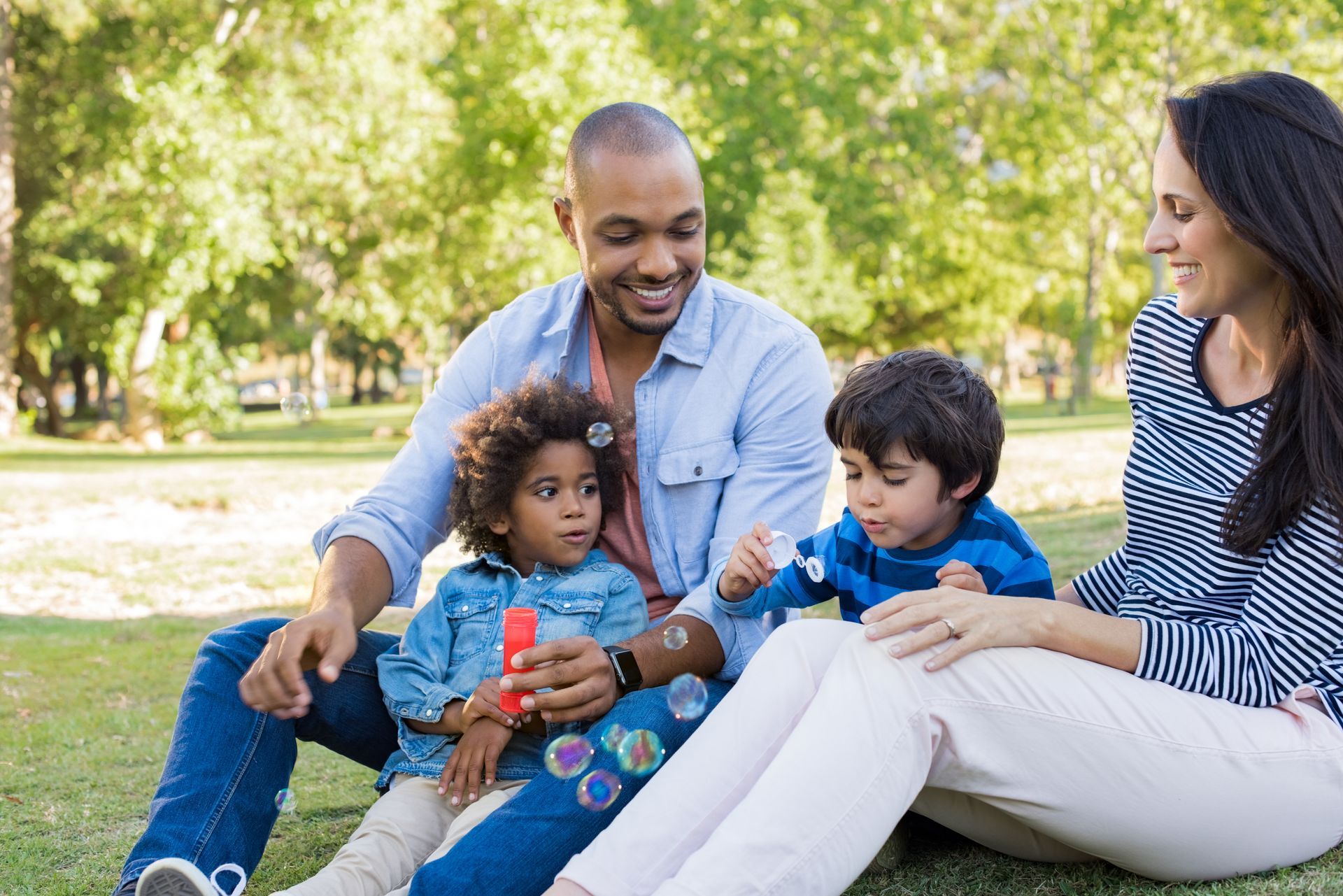 Family in a park blowing bubbles: father with two children, mother, seated on grass, sunny day.