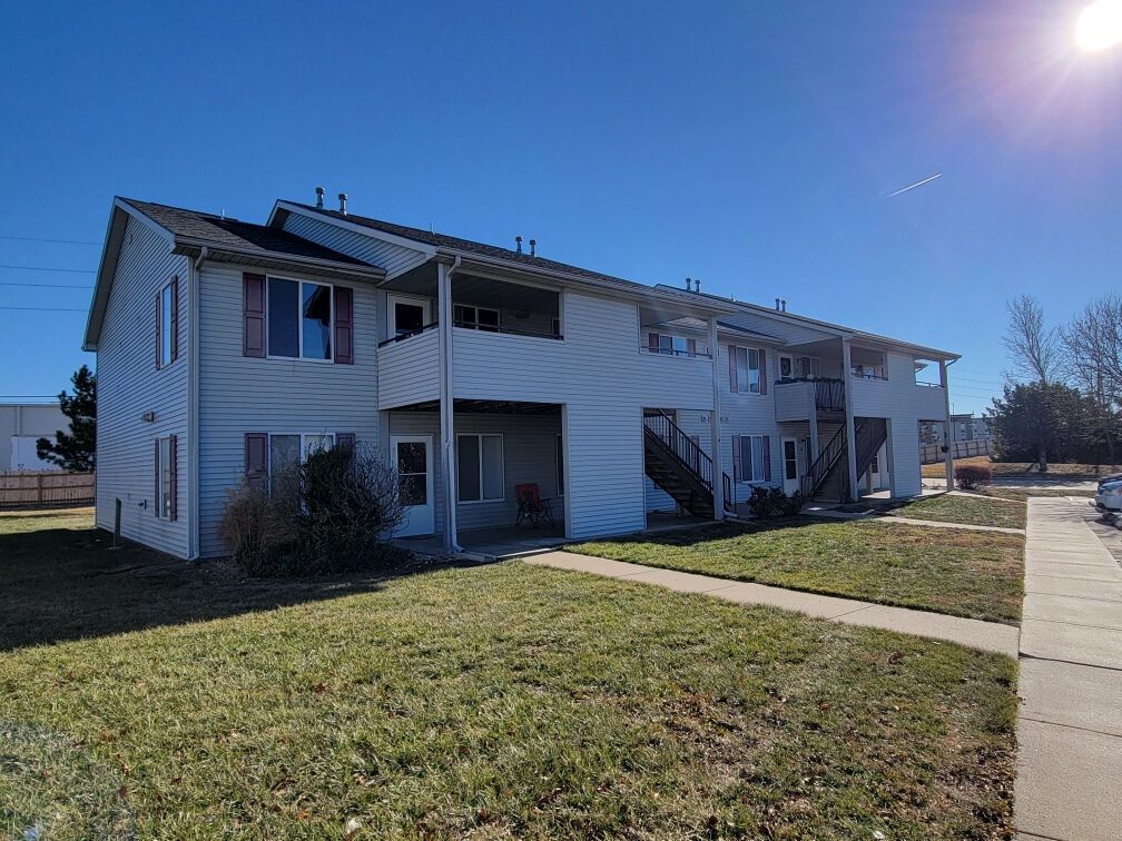 Two-story apartment building with light-colored siding and brown shutters on a sunny day. Green grass and a sidewalk in the foreground.