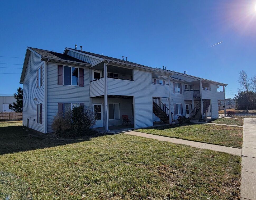 Two-story apartment building with light siding, brown stairs, and a pathway in front under a clear blue sky.