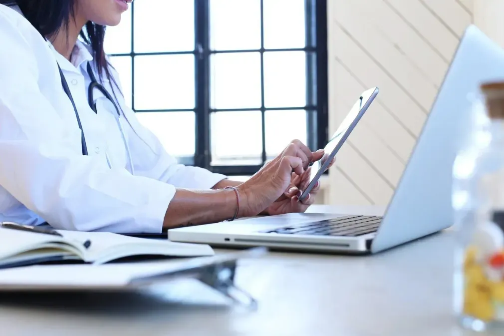 A doctor is sitting at a desk using a laptop and a tablet.