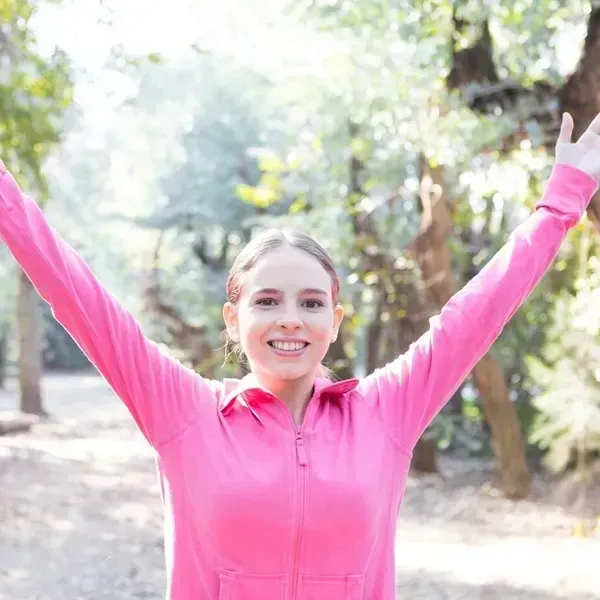 A woman in a pink jacket is standing in the woods with her arms outstretched.