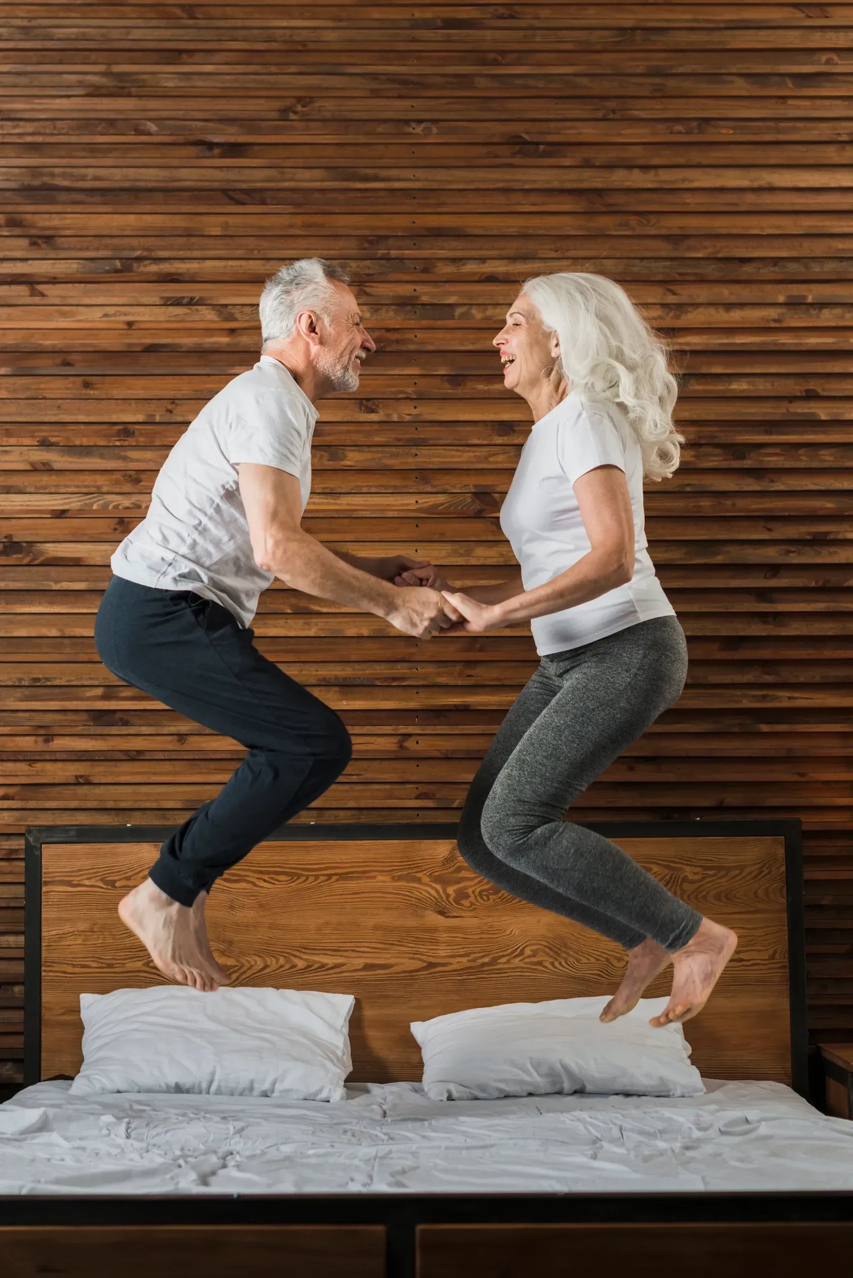 A man and a woman are jumping on a bed holding hands.