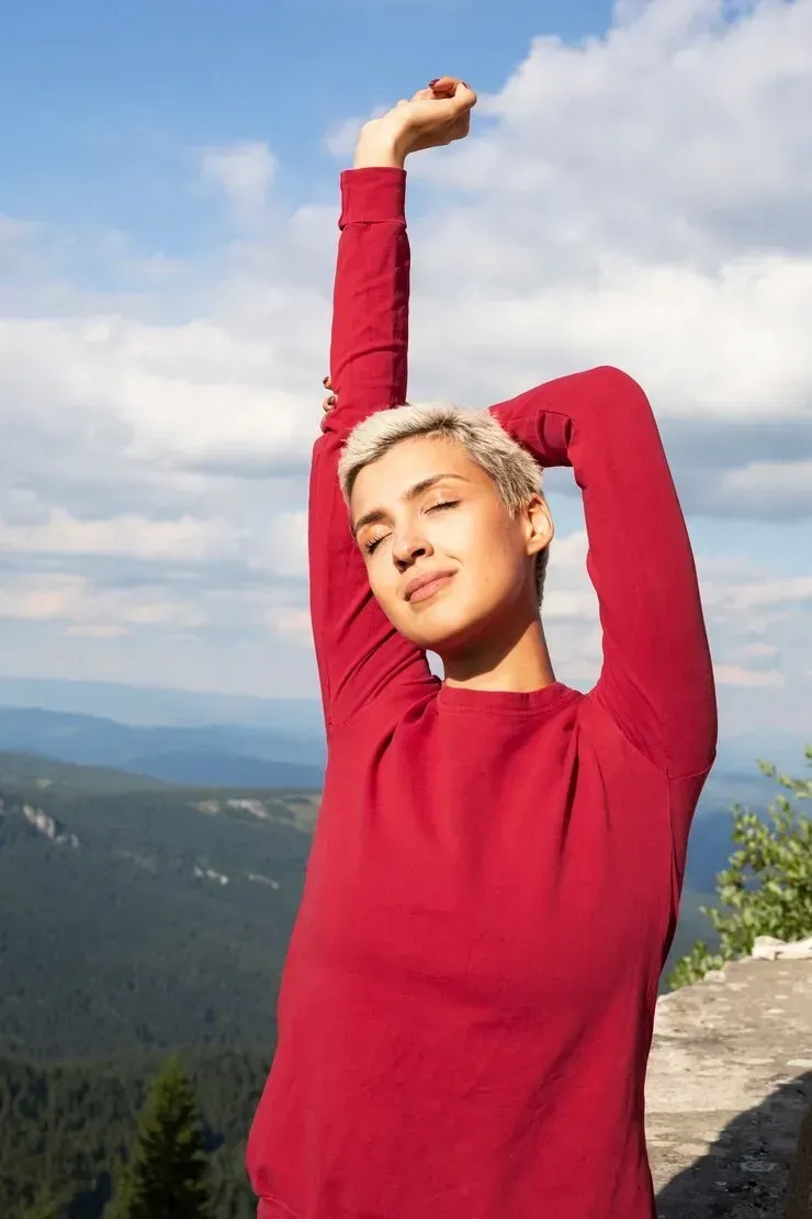 A woman in a red sweater is stretching her arms in front of a mountain.