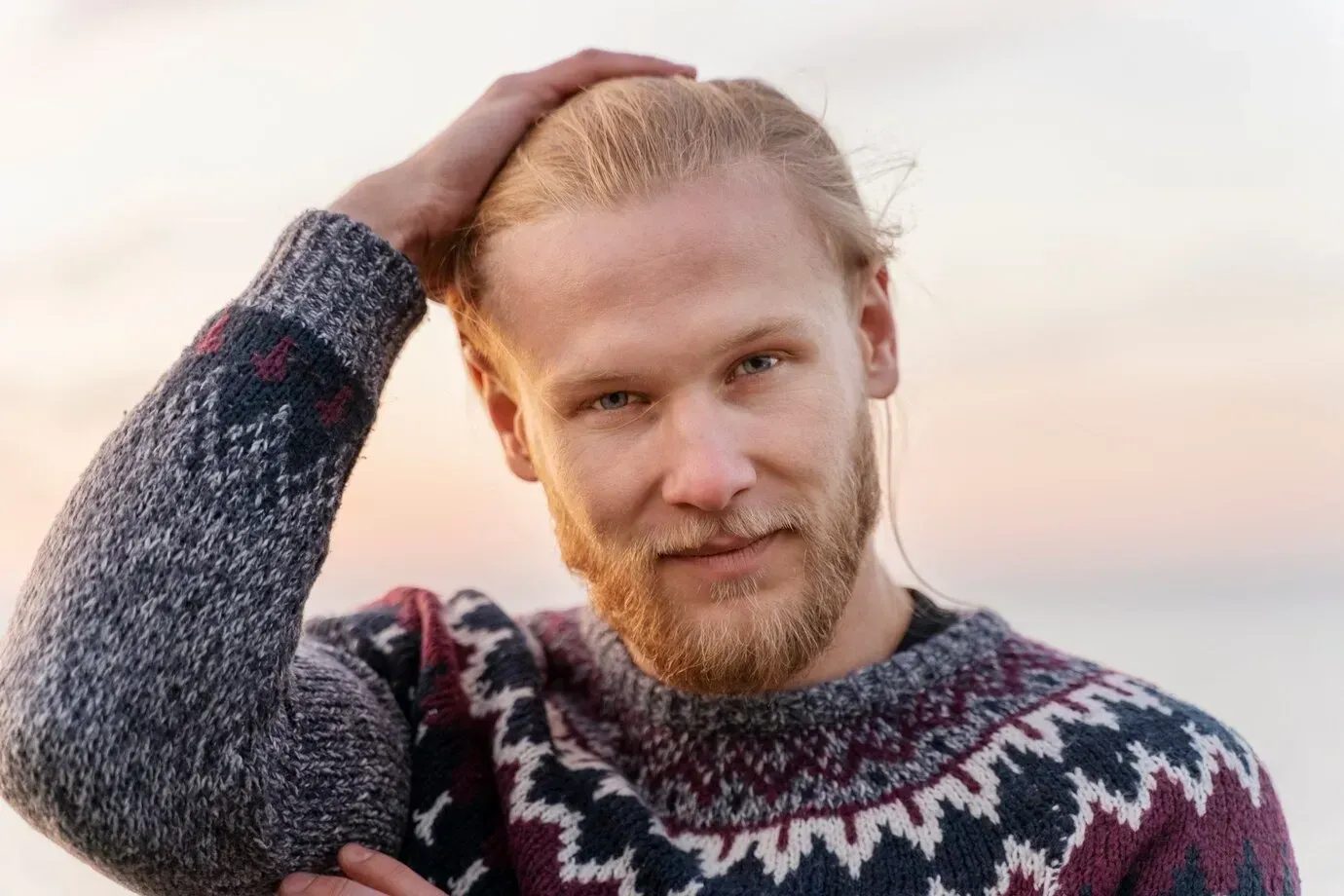 A man with a beard is wearing a sweater and touching his hair.
