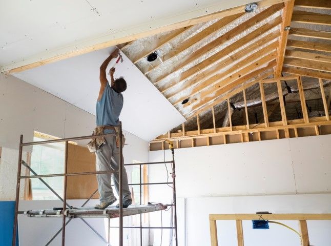 Construction worker installing drywall on a ceiling while standing on a scaffold in a room under renovation.